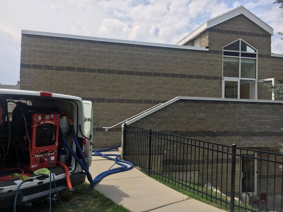 Carpet Cleaning Services in Salt Lake City - Father and Son Carpet Cleaning A van offering Carpet Cleaning services, equipped with hoses and tools, is parked outside a modern brick building in Salt Lake City, with hoses stretching up the sidewalk toward the entrance.
