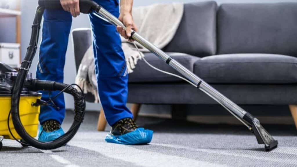 Carpet Cleaning and Home Services in Sandy, Utah - Father and Son Carpet Cleaning A person wearing blue shoe covers is vacuuming a carpet with a commercial vacuum cleaner in a Sandy Utah living room, highlighting professional carpet cleaning and home services near a grey sofa.