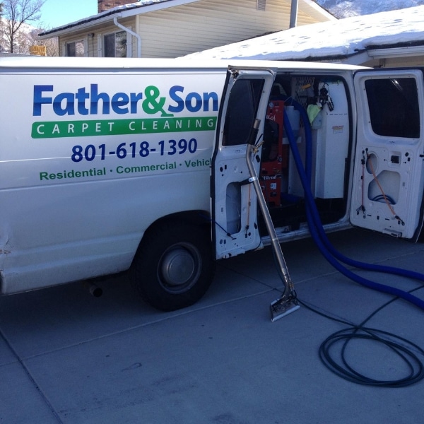 A white "Father & Son Carpet Cleaning" van, offering cleaning services in Salt Lake County, is parked in a driveway with its door open, displaying cleaning equipment and hoses extending onto the ground.