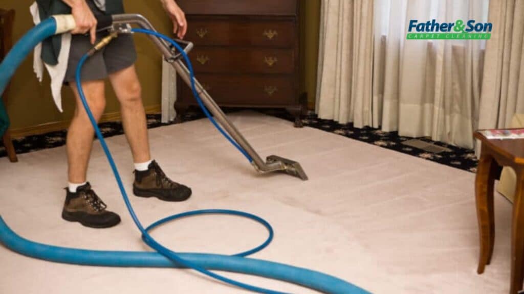 Why Carpet Stretching Matters More Than You Think - Father and Son Carpet Cleaning A person uses a carpet cleaning machine with a long blue hose to clean a beige carpet in a furnished room, highlighting the importance of regular flooring maintenance. The Father & Son Carpet Cleaning logo is visible in the top right corner.