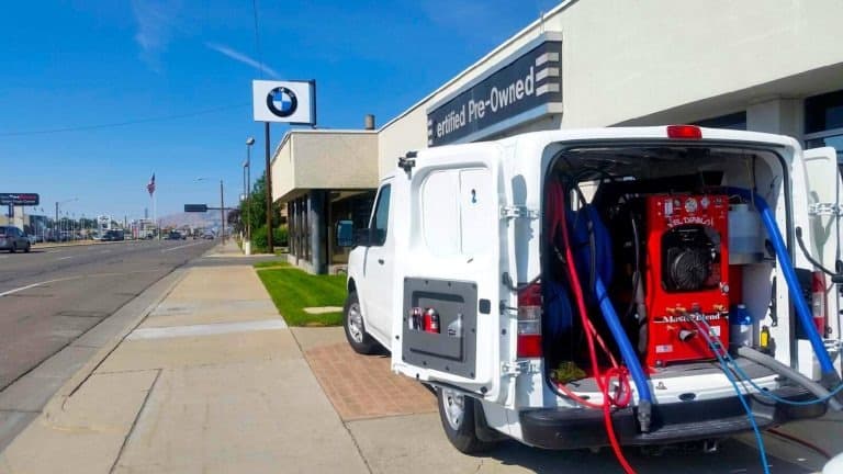 Professional Carpet Cleaning & Home Restoration in West Valley City, UT - Father and Son Carpet Cleaning A white van equipped with carpet cleaning equipment and hoses in the back is parked outside a BMW Certified Pre-Owned dealership on a city street in West Valley City, highlighting professional home restoration and cleaning services.