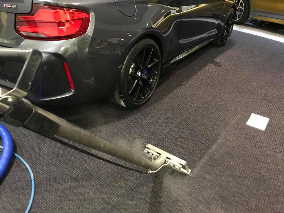 A professional carpet cleaning vacuum wand cleans a dark carpeted floor next to a parked gray car.